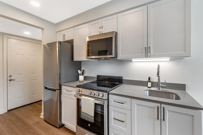 Renovated Package II kitchen with stainless steel appliances, light grey quartz countertops, white shaker cabinetry, and hard surface flooring - eaves Thousand Oaks