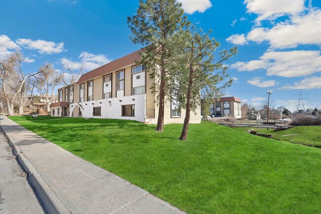 Charming apartment building surrounded by lush green lawns and picturesque trees under a vibrant blue sky. - Country Club