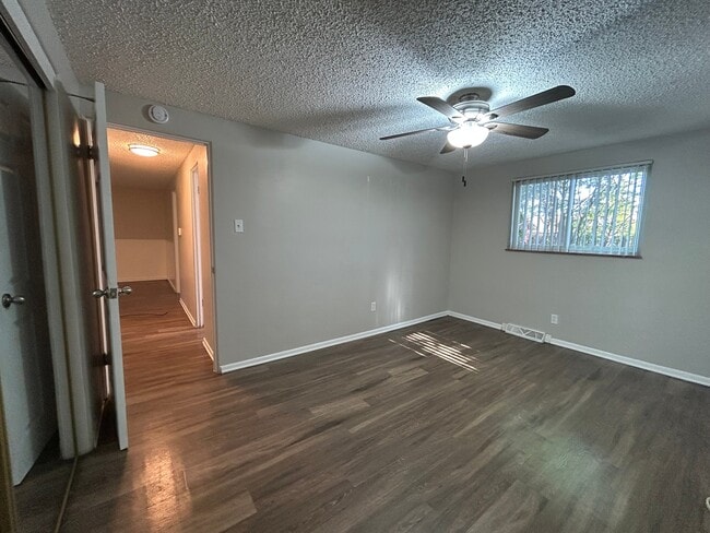 Inviting empty room with warm wood flooring, ample natural light from a window with blinds, and a stylish ceiling fan. - Boardwalk Apartments