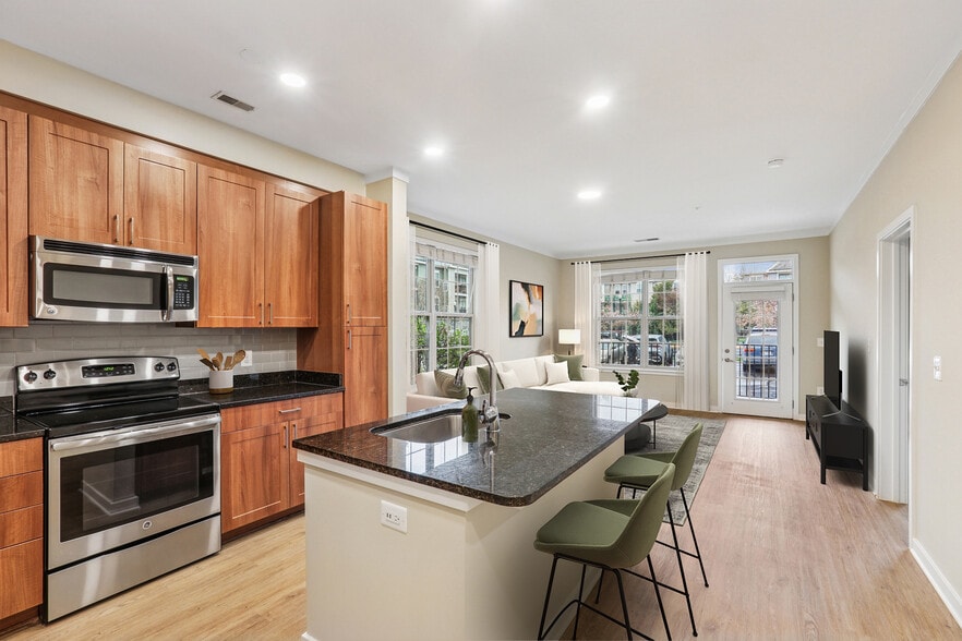 Renovated Package I kitchen with stainless steel appliances, oak cabinetry, grey tile backsplash, black granite countertops, and updated hard surface flooring and lighting - Avalon Falls Church