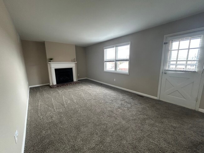 Bright and spacious living room with plush carpet, elegant fireplace, and abundant natural light from large windows. - Woods Mill Park Apartments & Townhomes