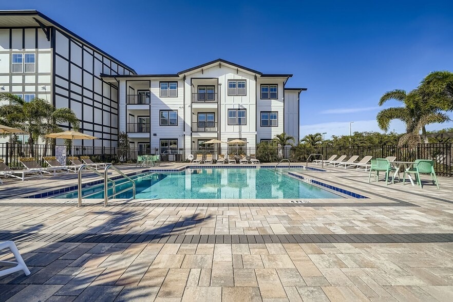 Luxurious apartment complex with a pristine outdoor pool area, surrounded by stylish loungers and palm trees under a clear blue sky. - Magnolia at Palm Bay