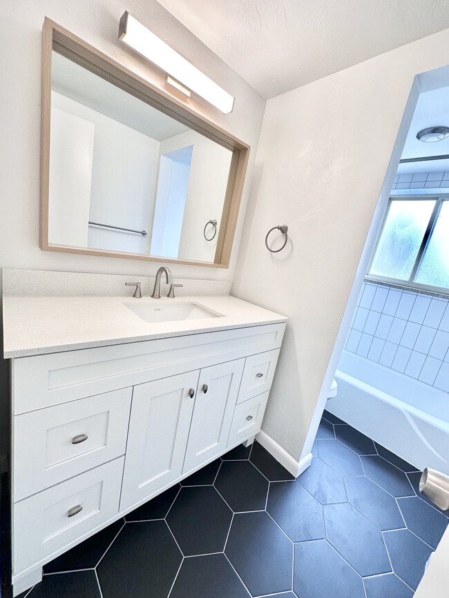 Bright and modern bathroom featuring sleek black hexagonal tiles, a stylish white vanity with ample storage, and a spacious bathtub area. - Columbine Plaza