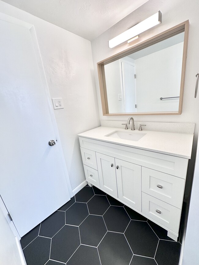 Modern bathroom with sleek white vanity, large mirror, and stylish hexagonal black tiles. - Columbine Plaza