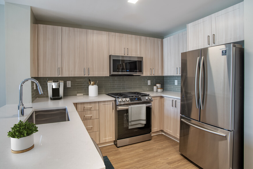 Renovated Package II kitchen with oak cabinetry, white quartz countertops, stainless steel appliances, grey title backsplash, and hard surface flooring - Avalon at Gallery Place