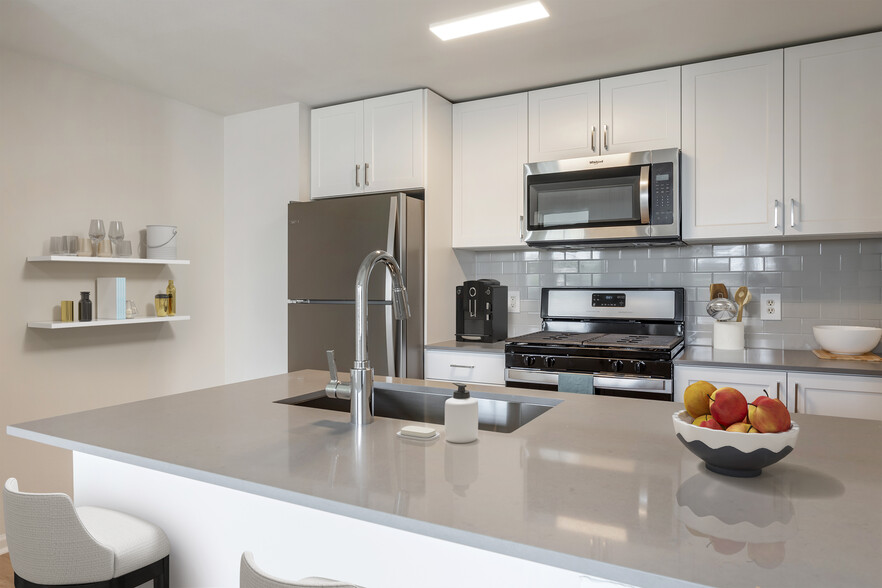 Finish Package II kitchen with white cabinetry, grey quartz countertops, extended hard surface flooring in select units, stainless steel appliances, and tile backsplash - Avalon Westbury