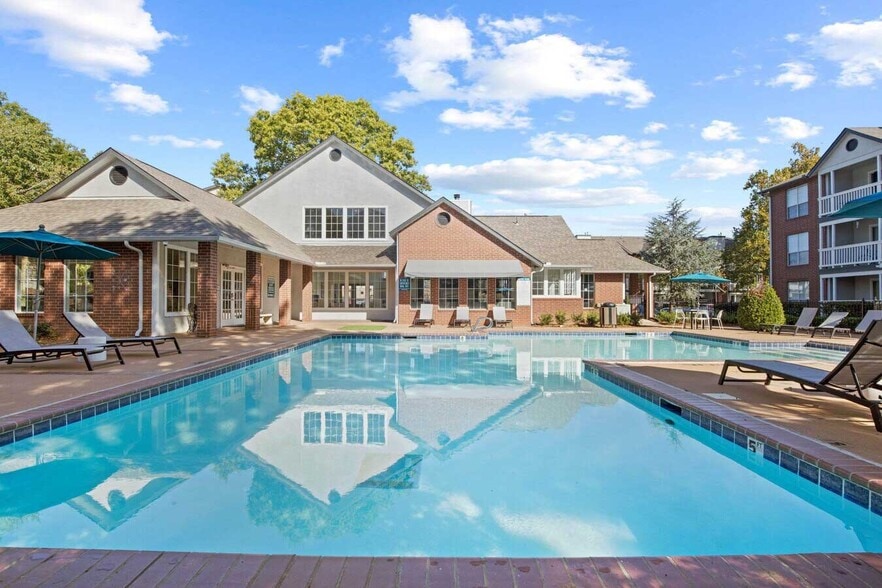 Community pool area featuring lounge chairs, umbrellas, and brick clubhouse under a bright blue sky with trees - Parc 1010
