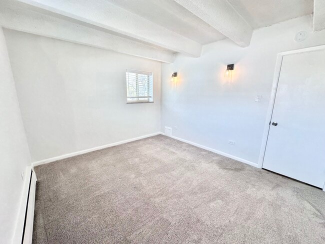 Bright and inviting empty room with plush carpet, white walls, and natural light from a small window. Elegant wall sconces add a modern touch. - Columbine Plaza