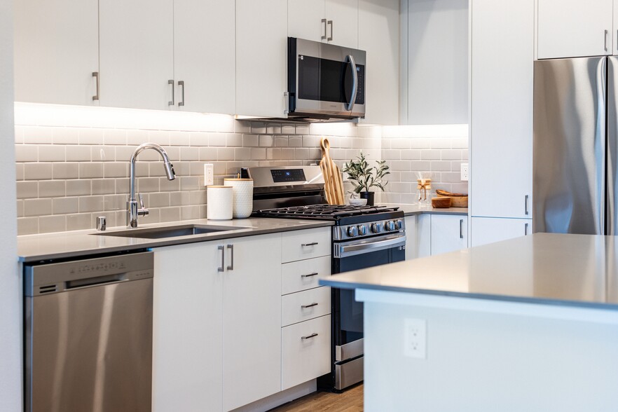 Kitchen with grey quartz countertops, white cabinetry, grey subway tile backsplash, stainless steel appliances, and hard surface flooring - Avalon Bothell Commons