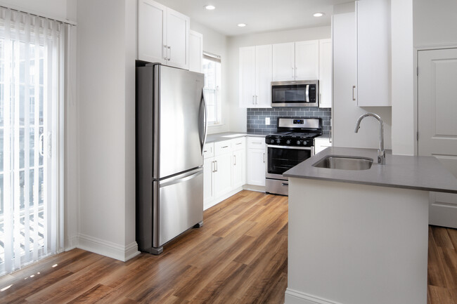 Kitchen with white cabinetry, grey quartz countertops, grey tile backsplash, stainless steel appliances, and hard surface flooring - Avalon at the Hingham Shipyard