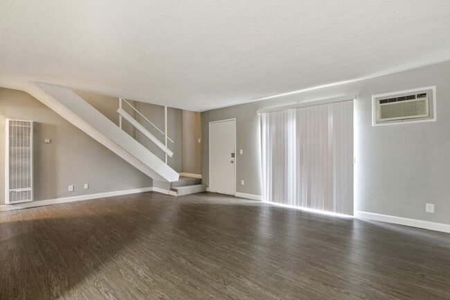Living room with air conditioning on the right, wood-style flooring, vertical blinds, carpeted staircase to the second floor - Lemar Gardens