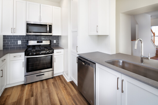 Kitchen with white cabinetry, grey quartz countertops, grey tile backsplash, stainless steel appliances, and hard surface flooring - Avalon at the Hingham Shipyard