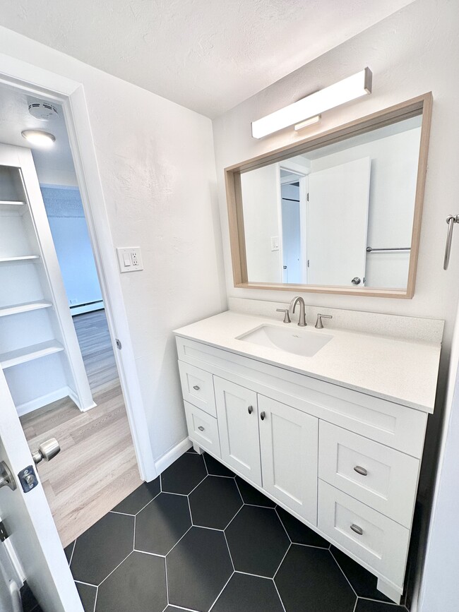 Modern bathroom featuring a sleek white vanity with ample storage, large mirror, and elegant black hexagon floor tiles. - Columbine Plaza