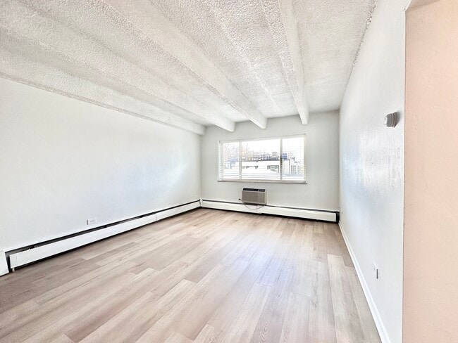Bright and airy living space with large window, wood flooring, and modern baseboard heating. - Columbine Plaza