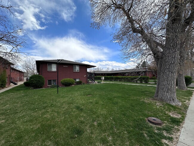 Charming brick apartment building nestled in a lush green yard with mature trees and a vibrant blue sky backdrop. - 7250-7290 Newton Street
