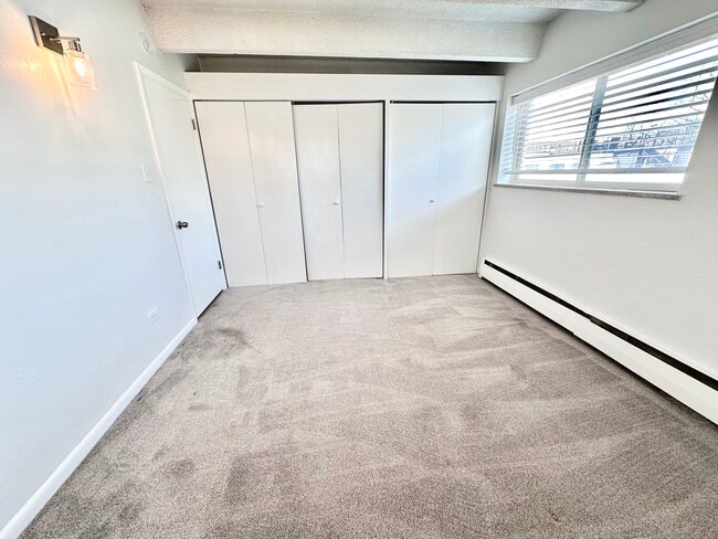 Bright, spacious bedroom with plush carpeting and large window featuring elegant blinds, enhancing natural light and offering a modern, cozy feel. - Columbine Plaza