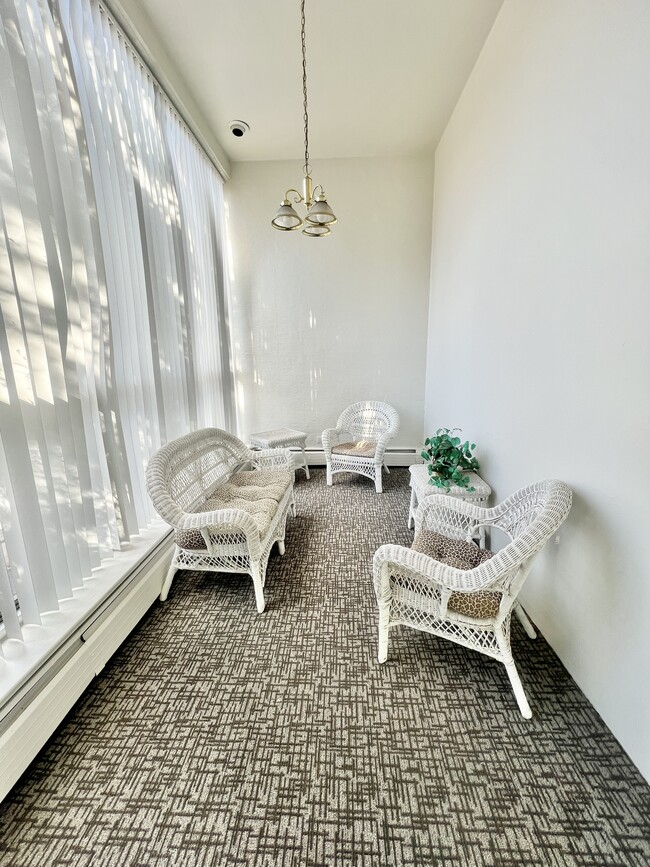 Charming sunroom with white wicker furniture and soft carpeting, basking in natural light. - Rhapsody Apartments