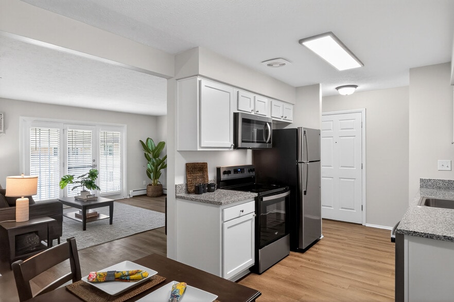 Modern open-plan kitchen and living area featuring sleek stainless steel appliances, elegant white cabinetry, and abundant natural light through large windows with blinds. - University Oaks