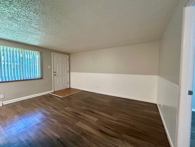Inviting living space with sleek wood flooring, large window with blinds, and modern entry door. - Boardwalk Apartments