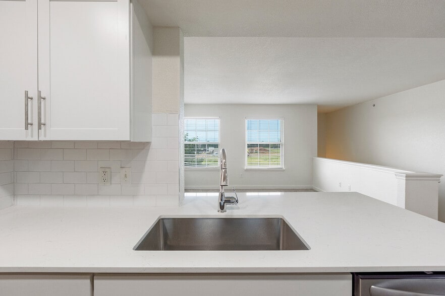 Bright modern kitchen with sleek white cabinetry, a spacious countertop, and stainless steel sink overlooking a sunlit living area with large windows. - Pointe at North Penn