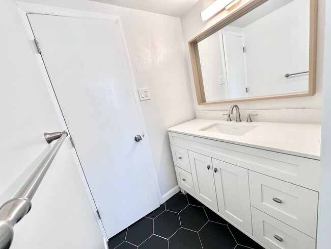 Sleek bathroom featuring a stylish vanity with a large mirror and hexagonal black tile flooring. - Columbine Plaza