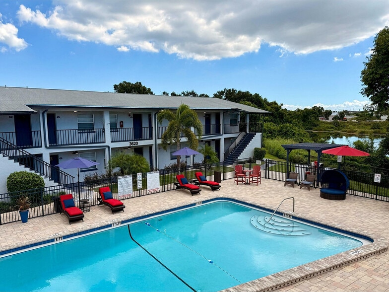 Charming apartment complex featuring a picturesque pool area with red lounge chairs, umbrellas, and lush greenery backdrop. - Waterside Village