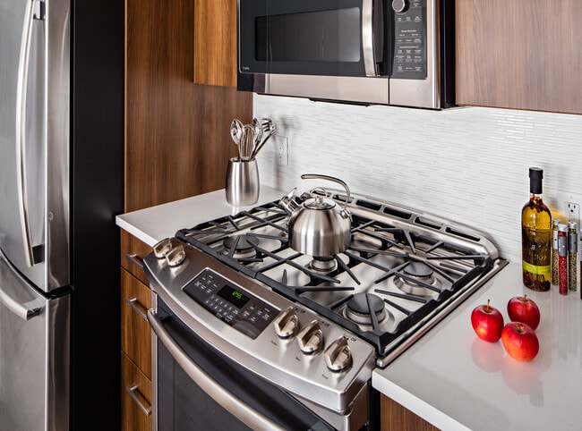 Signature Collection Kitchen with oak cabinetry, light grey quartz countertops, white tile backsplash, upgraded stainless steel appliances, and hard surface plank flooring - Avalon Princeton