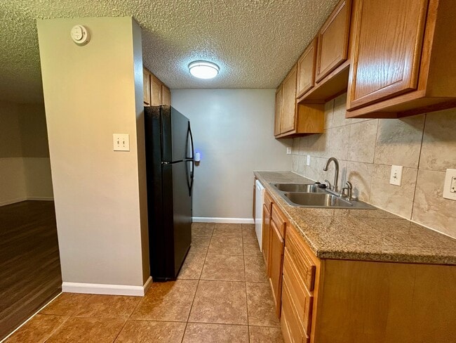 Modern kitchen featuring granite countertops, dual sink, wooden cabinetry, and sleek black refrigerator with tile flooring. - Boardwalk Apartments