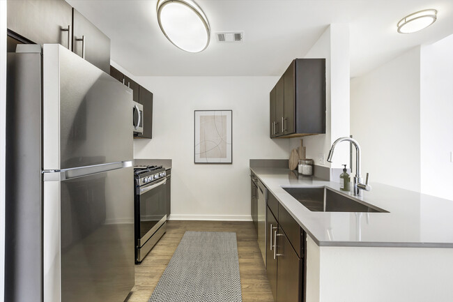 Renovated Package I kitchen with stainless steel appliances, grey quartz countertops, dark brown cabinetry, white tile backsplash, and hard surface flooring - Avalon at Wesmont Station