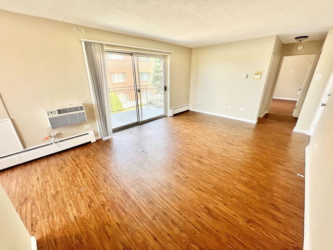Bright living room with elegant wood flooring, large sliding glass doors, and modern wall-mounted AC unit, leading to a welcoming balcony. - Bonneville Apts