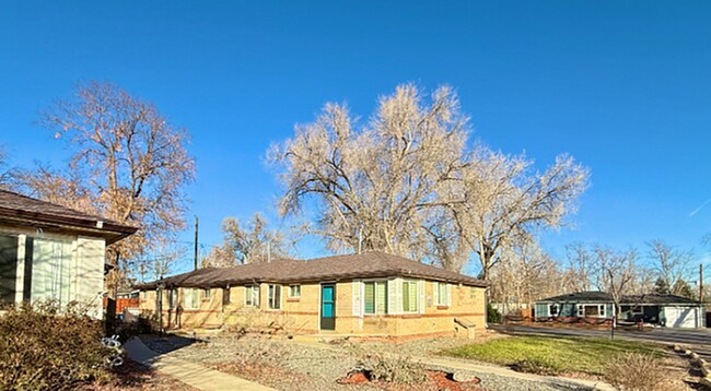 Charming single-story brick home with large windows, surrounded by mature trees and set against a bright blue sky. - Flats at 16th