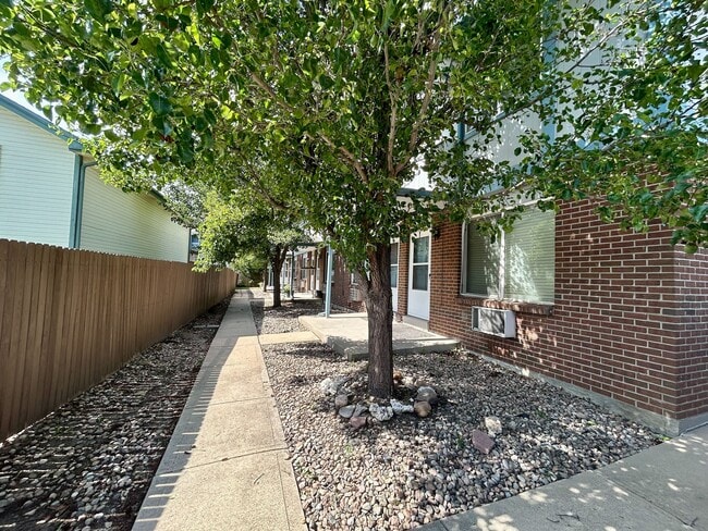 Charming walkway lined with lush green trees leading to a brick apartment building, offering a serene and inviting residential atmosphere. - 10756 West 12th Lane